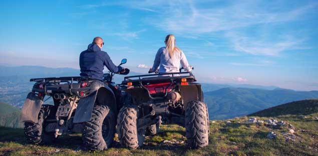 A man and a woman sit on their ATVs at the top of a mountain, looking out over a vast valley and rolling hills under a clear blue sky - labonteins.com