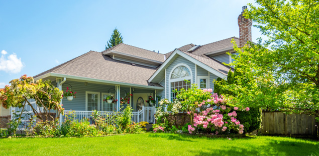 A charming two-story blue-grey suburban home with a white-railed porch, featuring a lush green lawn and vibrant pink rhododendrons in the foreground -labonteins.com