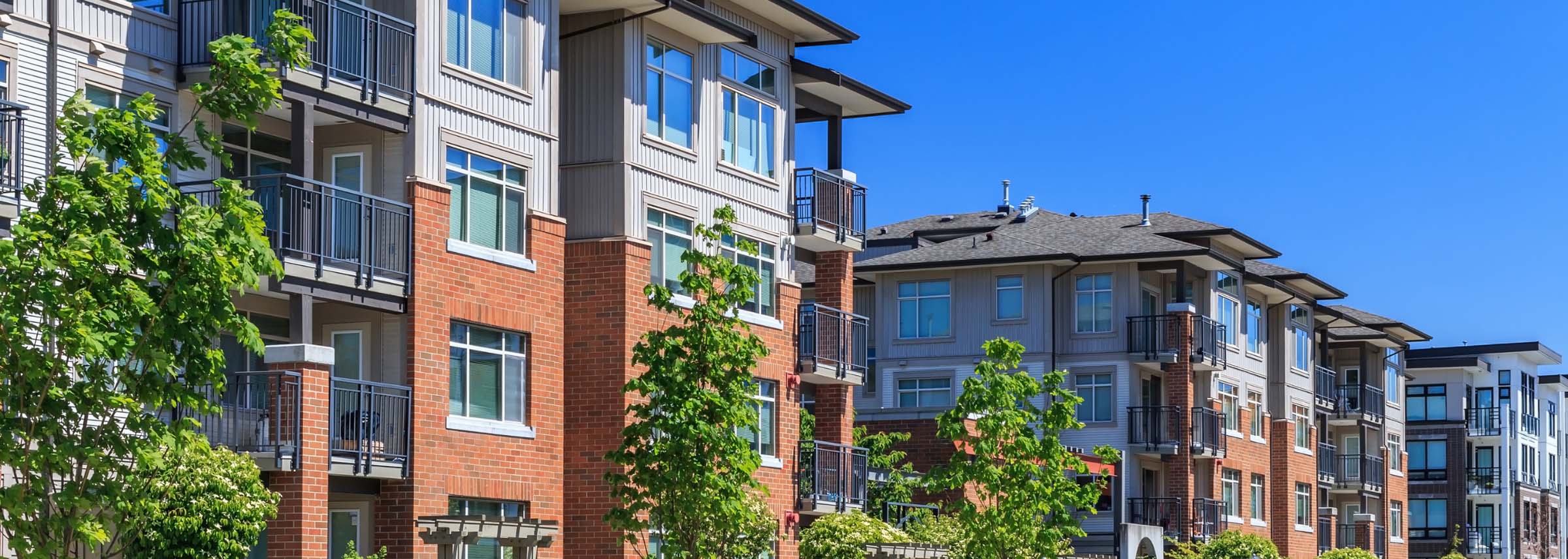 A row of modern multi-story apartment buildings with brick and siding facades and dark metal balconies under a clear blue sky- labonteins.com