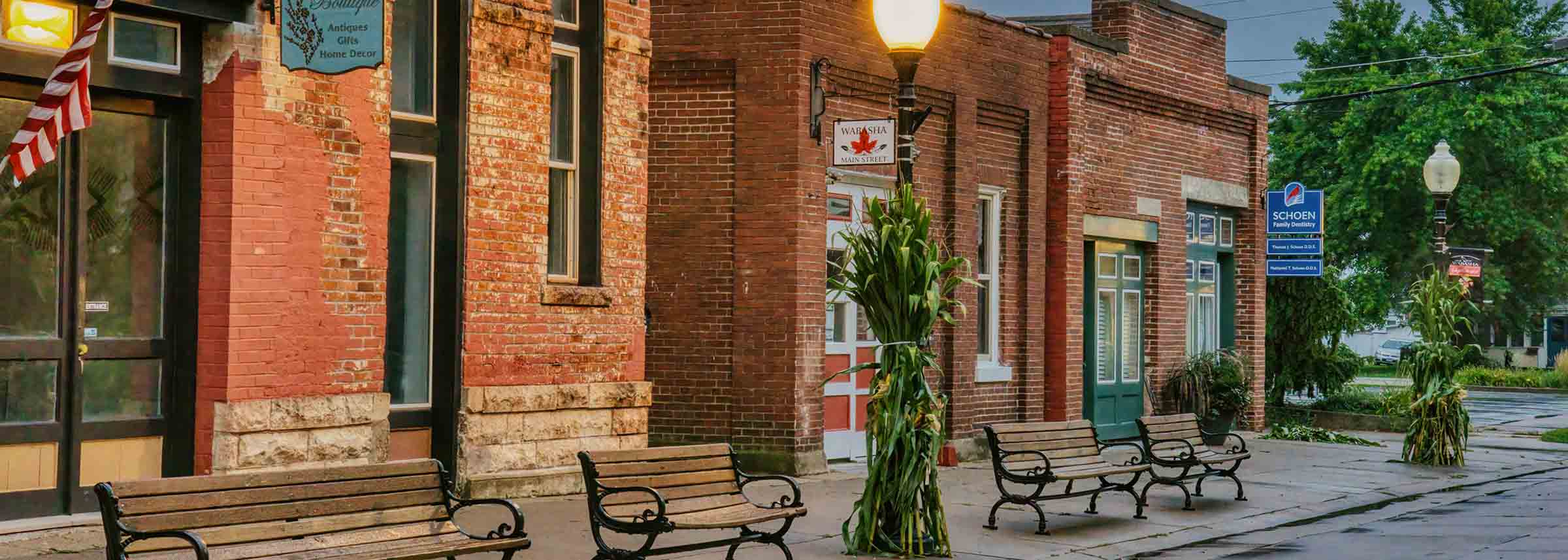 A row of red brick buildings along a wet sidewalk, featuring wooden park benches and seasonal corn stalk decorations on lampposts - labonteins.com