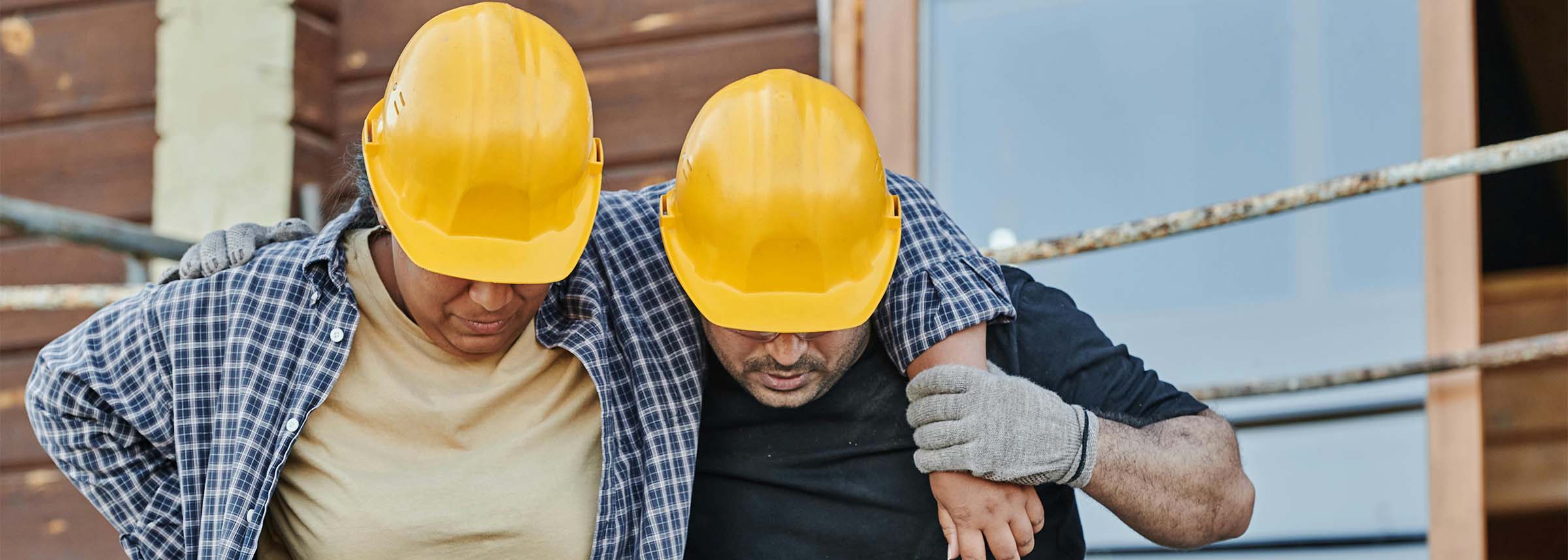 A construction worker in a yellow hard hat clutches their lower back in pain while being supported by a colleague at a job site -labonteins.com